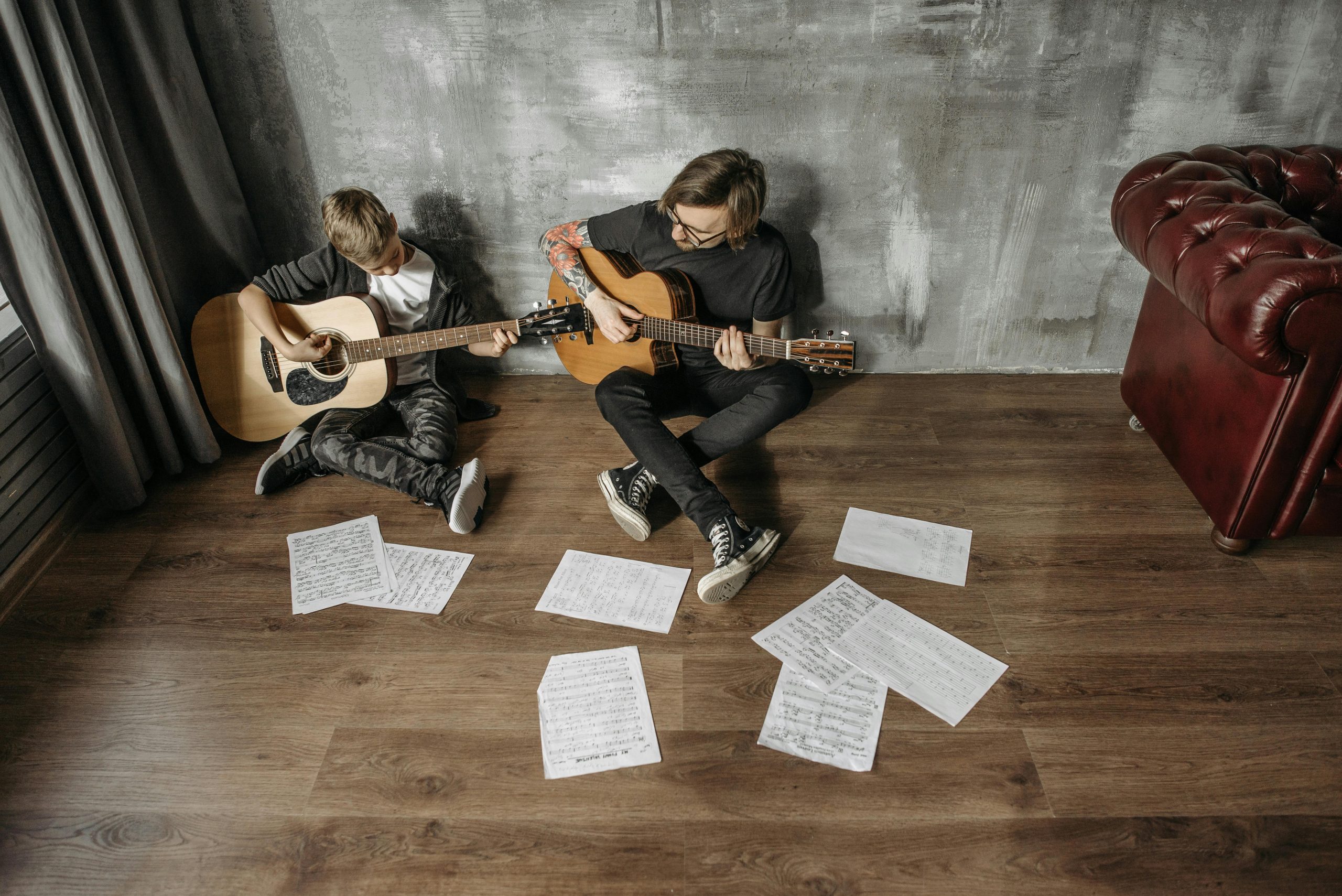 Adult and teenager practicing guitars with sheet music spread across a wooden floor.